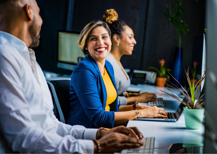 três colaboradores(duas mulheres e um homem) trabalhando e sorrindo no escritório